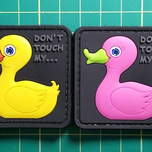 Può includere: Due toppe nere con un design a forma di papera di gomma. La toppa a sinistra presenta una papera gialla con un becco arancione e il testo "Don't Touch My..." La toppa a destra presenta una papera rosa con un becco verde e lo stesso testo.