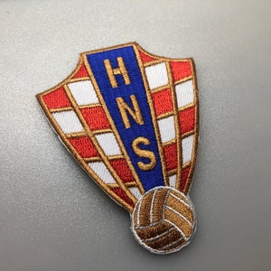 May include: A red, white, and blue embroidered patch with the letters "HNS" in a blue rectangle. The patch is shaped like a shield and has a gold embroidered soccer ball at the bottom.