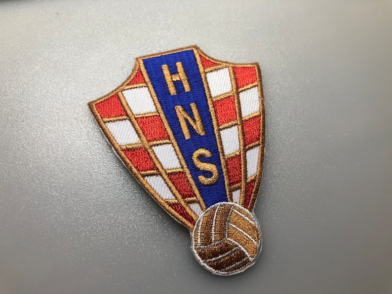 May include: A red, white, and blue embroidered patch with the letters "HNS" in a blue rectangle. The patch is shaped like a shield and has a gold embroidered soccer ball at the bottom.