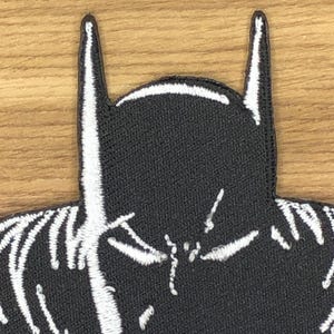 Puede incluir: Parche bordado en blanco y negro con la silueta de una máscara de superhéroe. El parche tiene un fondo negro con detalles bordados en blanco, incluyendo las orejas puntiagudas y rasgos faciales distintivos. Ideal para ropa o accesorios.