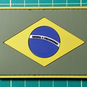 以下が含まれることがあります： ブラジルの国旗が描かれた緑と黄色の布製パッチ。国旗には、白い文字で「ORDEM E PROGRESSO」と書かれた青い円が描かれています。