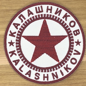 Può includere: Toppa circolare con centro bianco e bordo bordeaux. Al centro è presente una stella bordeaux. Le parole "KALASHNIKOV" sono ricamate in bordeaux attorno alla stella. La toppa ha un aspetto strutturato.