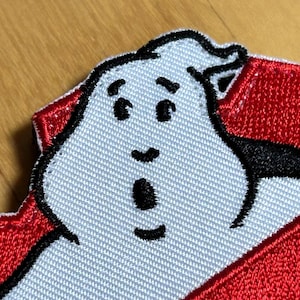 Pode incluir: Etiqueta bordada com um fantasma branco com contorno preto sobre um fundo vermelho. O fantasma tem uma expressão de surpresa com a boca aberta e olhos grandes. O fundo vermelho tem a forma de uma borda irregular.