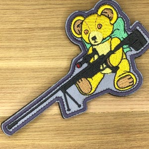 May include: Embroidered patch featuring a yellow teddy bear with a green bow, holding a black rifle. The patch has a gray background with a purple border. The bear has brown accents and a red detail on the rifle.