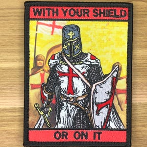 May include: Embroidered patch depicting a knight in armour, holding a sword and shield with a red cross. The patch includes the text "WITH YOUR SHIELD" at the top and "OR ON IT" at the bottom, set against a yellow background.