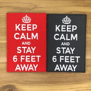 Puede incluir: Dos parches rectangulares, uno rojo y otro negro, con texto blanco y un gráfico de corona. El texto en cada parche dice "KEEP CALM AND STAY 6 FEET AWAY". Los parches están sobre una superficie de madera clara.