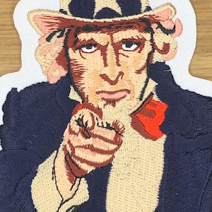 Puede incluir: Parche bordado que representa al Tío Sam señalando. El parche tiene un borde blanco, con el Tío Sam vistiendo un abrigo azul marino, una pajarita roja y un sombrero con estrellas.