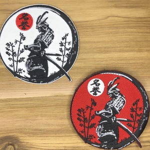 Könnte beinhalten: Zwei runde, gestickte Aufnäher mit einem Samurai-Krieger. Einer hat einen weißen Hintergrund mit roter Sonne und schwarzen Details. Der andere hat einen roten Hintergrund mit weißer Sonne und schwarzen Details. Beide Aufnäher haben japanischen Text.
