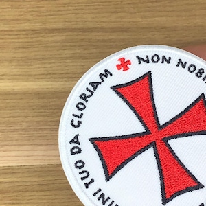 May include: A circular embroidered patch featuring a white background with a large red cross in the centre. The text "NON NOBIS DOMINE SED NOMINI TUO DA GLORIAM" is stitched in black around the border, accompanied by a small red cross symbol.