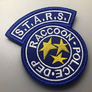Peut inclure: Un écusson brodé bleu et blanc avec le texte "S.T.A.R.S. Raccoon Police Dept." et trois étoiles jaunes.