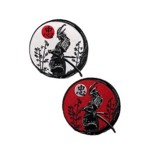 May include: Two circular embroidered patches depicting a samurai warrior. One has a white background with a red sun and black detailing. The other has a red background with a white sun and black detailing. Both patches feature Japanese characters.