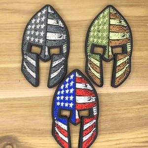 May include: Three embroidered patches in the shape of a Spartan helmet. One patch features the American flag in red, white, and blue. Another has a gray and white flag design. The third patch has a camouflage pattern.