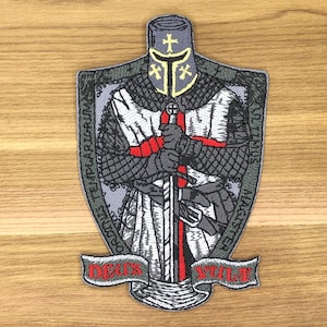 May include: Embroidered patch featuring a medieval knight in full armor, holding a sword with a red cross. The design includes a shield shape with text around the border and a banner at the bottom that reads "DEUS VULT". The color palette includes gray, white, red, and gold.
