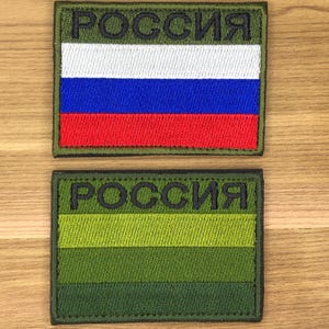 May include: Two embroidered patches. The top patch features the Russian flag with white, blue, and red horizontal stripes, and the word "РОССИЯ" in green. The bottom patch has green and olive stripes with the same text.