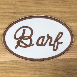 Op de afbeelding: Een ovaalvormige patch met een witte kern en een bruine rand. Het woord "Barf" is in bruin cursiefschrift over het midden geborduurd. De patch is ontworpen om op kleding of accessoires te worden genaaid.