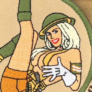 Puede incluir: Parche bordado que representa a una mujer rubia con un sombrero verde y ropa naranja. El parche tiene un fondo beige y un borde verde. La mujer lleva guantes blancos y tiene una sonrisa roja.