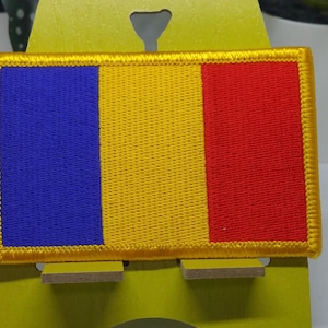 May include: A blue, yellow, and red embroidered patch with a yellow border. The patch is on a yellow stand.
