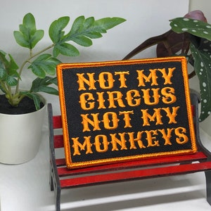 May include: A black embroidered patch with orange lettering that reads "Not My Circus Not My Monkeys". The patch is displayed on a small wooden bench.