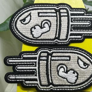 May include: Two embroidered patches depicting a cartoon bullet with a grumpy face and a flexing arm. The patches are grey with black outlines and white accents, and are likely iron-on or sew-on.