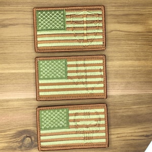 Puede incluir: Tres parches bordados con un diseño de bandera estadounidense. Los parches tienen un campo de estrellas verde y rayas beige y blancas. Cada parche presenta un emblema diferente: un escudo, una calavera y una figura estilizada.