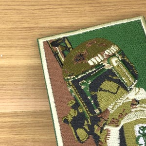 Puede incluir: Parche bordado con una imagen detallada del casco y la armadura de un personaje. El parche tiene un fondo verde con bordes marrones y blancos. El casco del personaje es principalmente verde oliva y marrón.