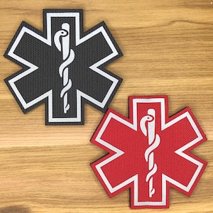 May include: Two embroidered Star of Life patches. One is black with a white border and a white staff of Asclepius. The other is red with a white border and a white staff of Asclepius. Both are on a wooden surface.