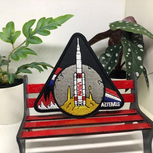 Puede incluir: Un parche triangular negro con un cohete blanco y rojo en el centro. El cohete est&aacute; sobre una luna gris con una llama amarilla y naranja. El parche tiene las palabras "NASA" y "ARTEMIS" en &eacute;l.