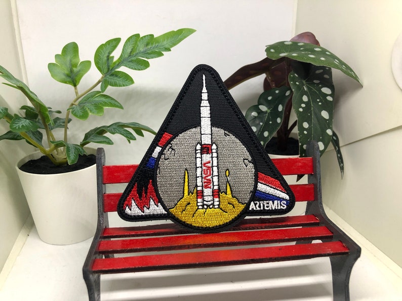 Puede incluir: Un parche triangular negro con un cohete blanco y rojo en el centro. El cohete est&aacute; sobre una luna gris con una llama amarilla y naranja. El parche tiene las palabras "NASA" y "ARTEMIS" en &eacute;l.