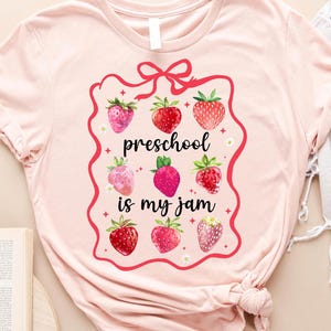 May include: Heather peach t-shirt with a strawberry design. The shirt features a red frame with the text "preschool is my jam" and illustrations of strawberries. The shirt is part of a preschool or teacher-themed collection.