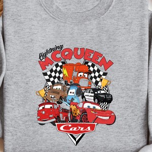 a gray sweatshirt with a graphic of Lightning McQueen and various cars from the Cars movie, along with a pair of jeans and sunglasses.