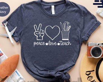 May include: A navy blue t-shirt with a white peace sign, heart, and pencil cup graphic. The text "peace love teach" is printed below the graphic.