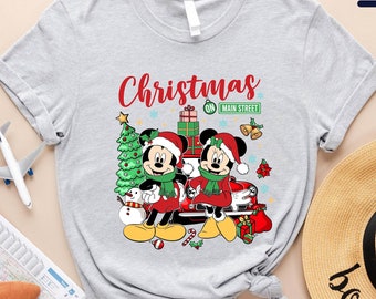 May include: Light gray t-shirt with a Christmas theme featuring Mickey Mouse and Minnie Mouse in front of a Christmas tree and a sign that says "Christmas on Main Street".