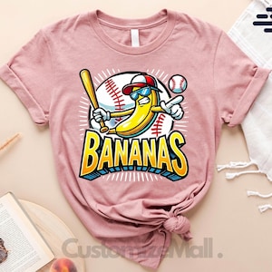 K&ouml;nnte beinhalten: Hellmauvefarbenes T-Shirt mit einer Comic-Banane, die eine Sonnenbrille und eine Baseballkappe tr&auml;gt, einen Schl&auml;ger h&auml;lt und zeigt. Das Wort "BANANAS" ist in einer blauen und gelben 3D-Schriftart gehalten. Ein Baseball und Baseballn&auml;hte sind im Hintergrund.