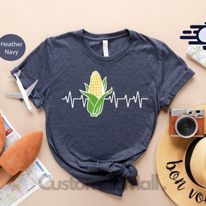 May include: A navy blue t-shirt with a white outline of a corn cob with a green husk and a white heartbeat line. The text "Next Day Shipping" is visible in the background.