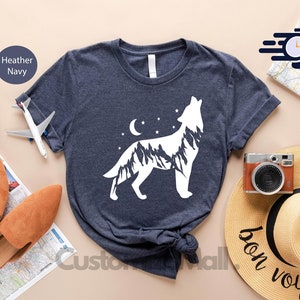 K&ouml;nnte beinhalten: Dunkelblau meliertes T-Shirt mit einem wei&szlig;en Wolfsdesign, Mond und Sternen. Das Bild zeigt Reiseaccessoires, eine Karte, einen Reisepass und einen Strohhut mit der Aufschrift "bon voyage".