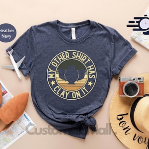 May include: A navy blue t-shirt with a vintage-style graphic that says "My Other Shirt Has Clay On It". The graphic features a clay pot with a plant inside and a yellow star on each side. The shirt is laid out on a table with other items, including a straw hat, a passport, and a pair of brown sandals.