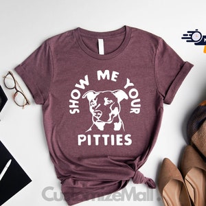May include: A maroon t-shirt with the text "Show Me Your Pitties" in white lettering. A white silhouette of a pit bull dog is centered in the text.