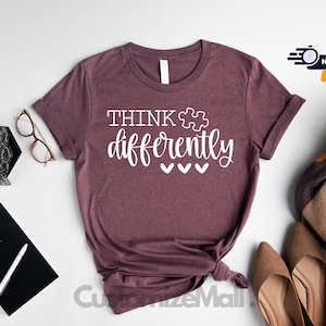 May include: A heather maroon t-shirt with the text "THINK differently" in white lettering. A white puzzle piece symbol is included in the text. Three white hearts are below the text.