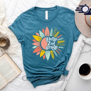 May include: Teal t-shirt with a sunflower design and the text "Hello, 2nd grade." The sunflower has yellow, orange, and coral petals. Pencils and rulers surround the text. The shirt is on a white surface with a hat and coffee cup.
