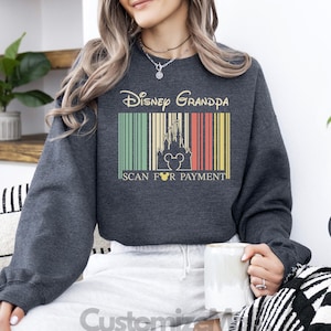 A woman wearing a gray sweatshirt with a Disney Grandpa design is sitting on a couch, holding a mug, and surrounded by potted plants.