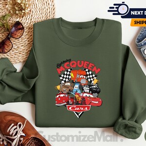 a green sweatshirt with a graphic of the Disney Cars characters Lightning McQueen and various cars, along with a pair of sunglasses and a pair of jeans.