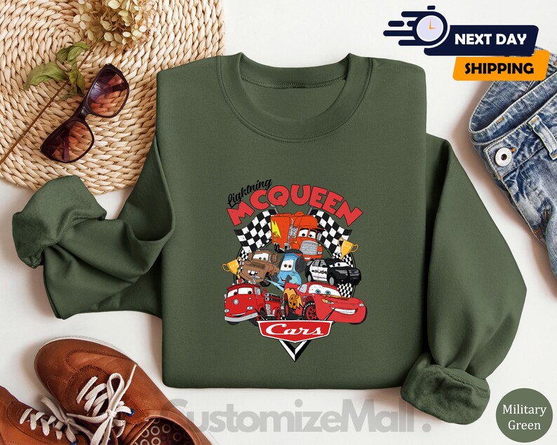 a green sweatshirt with a graphic of the Disney Cars characters Lightning McQueen and various cars, along with a pair of sunglasses and a pair of jeans.