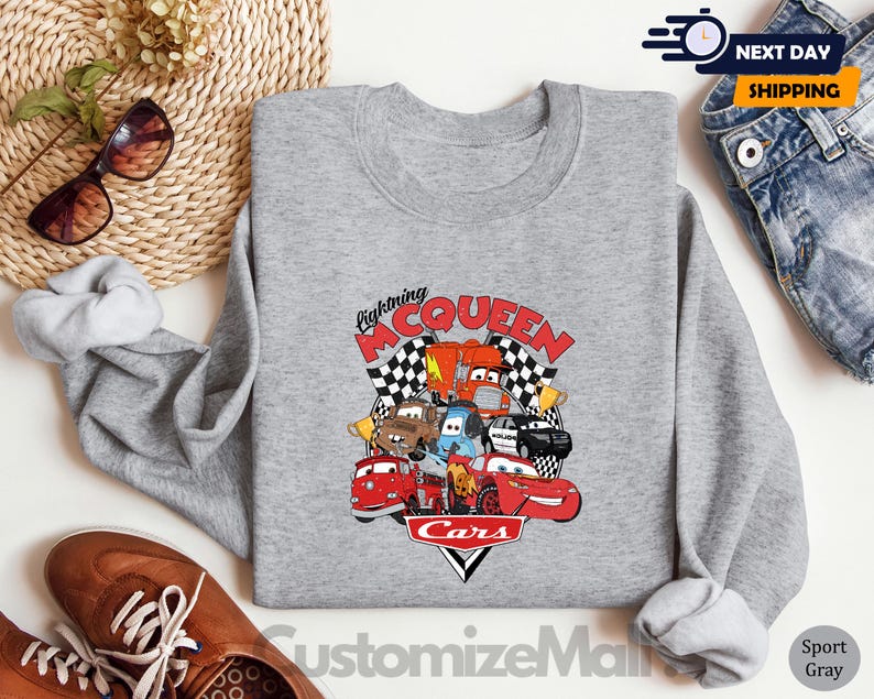 a gray sweatshirt with a graphic of Lightning McQueen and various cars from the Cars movie, along with a pair of jeans and sunglasses.
