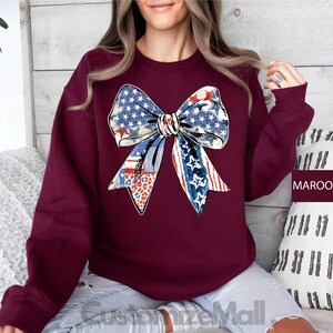 May include: A maroon sweatshirt with a large, colorful bow made of fabric with a red, white, and blue pattern. The bow is centered on the front of the sweatshirt.
