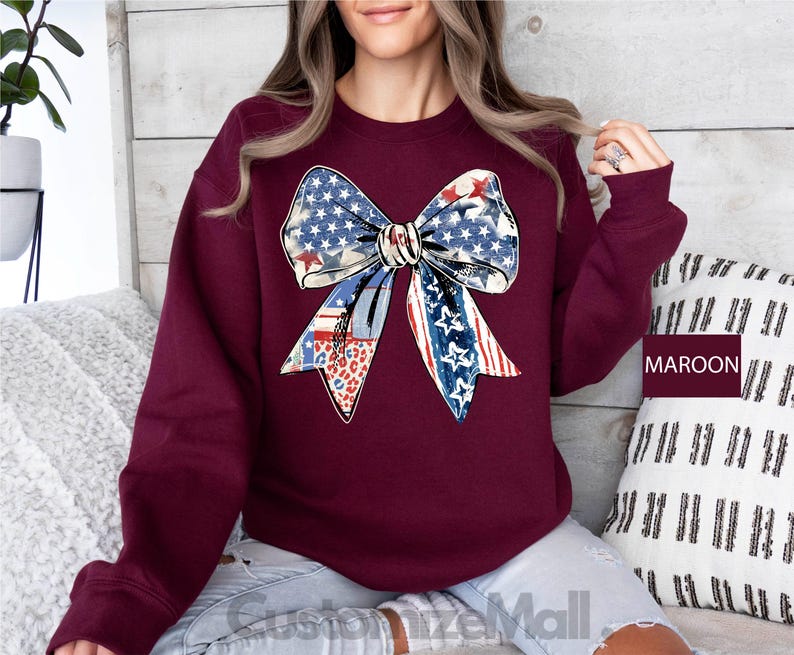 May include: A maroon sweatshirt with a large, colorful bow made of fabric with a red, white, and blue pattern. The bow is centered on the front of the sweatshirt.
