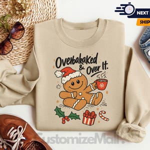 May include: Sand-colored sweatshirt with a gingerbread man graphic wearing a Santa hat and holding a mug. The text "Overbaked & Over It" is above the gingerbread man. Christmas-themed design with holly, a gift, and a candy cane.