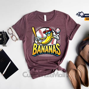 K&ouml;nnte beinhalten: Weinrotes T-Shirt mit einer Comic-Banane, die eine Sonnenbrille und eine Baseballkappe tr&auml;gt und einen Schl&auml;ger h&auml;lt, mit dem Wort "BANANAS" in Gelb und Blau. Das Shirt ist mit Accessoires versehen.