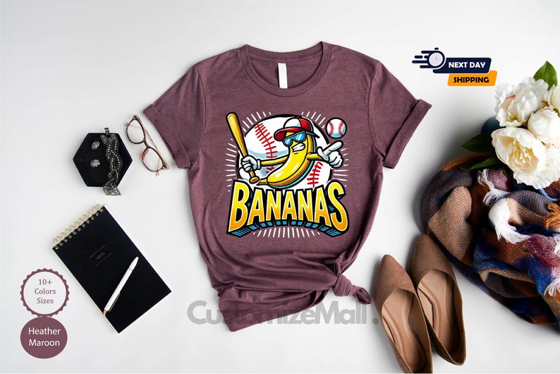 K&ouml;nnte beinhalten: Weinrotes T-Shirt mit einer Comic-Banane, die eine Sonnenbrille und eine Baseballkappe tr&auml;gt und einen Schl&auml;ger h&auml;lt, mit dem Wort "BANANAS" in Gelb und Blau. Das Shirt ist mit Accessoires versehen.