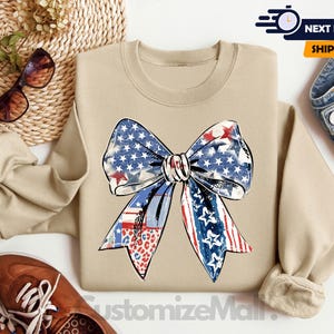 May include: A tan sweatshirt with a large red, white, and blue bow with stars and stripes. The bow is in a distressed, vintage style. The sweatshirt is a comfortable, casual style.