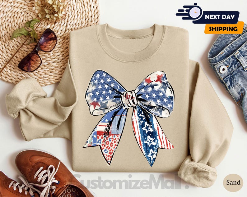 May include: A tan sweatshirt with a large red, white, and blue bow with stars and stripes. The bow is in a distressed, vintage style. The sweatshirt is a comfortable, casual style.